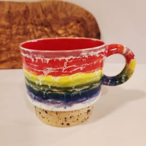 May include: A ceramic mug featuring a vibrant horizontal rainbow glaze pattern with red, yellow, green, blue, and purple bands. The interior is solid red, and the base is a speckled beige clay. A matching rainbow-glazed handle is attached to the side.