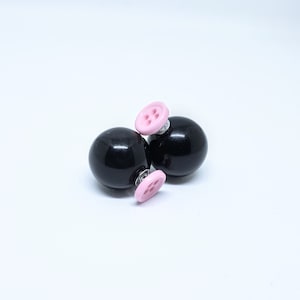 May include: A pair of black stud earrings with pink button accents. The earrings are round and have a shiny finish.