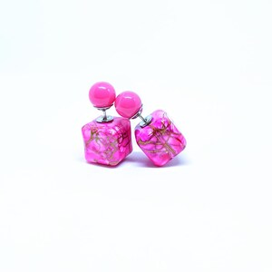 May include: A pair of pink double-sided earrings with a square shape and a pink ball on top. The earrings have a marbled design with gold accents.