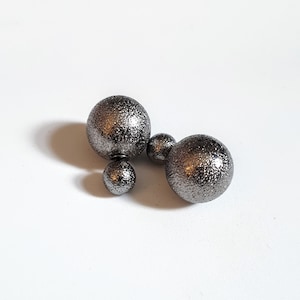 May include: A pair of dark grey double-sided stud earrings. Each earring has a textured, metallic finish with a large sphere at the front and a smaller sphere behind the ear. The earrings are on a white surface.