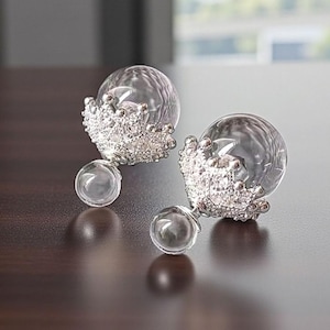 May include: A pair of clear, spherical earrings with a silver crown design. The earrings have a small, clear sphere at the base. The jewellery is displayed on a dark brown surface.
