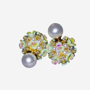 May include: A pair of floral stud earrings. Each earring features a large, pearlescent, round bead. The studs are adorned with small, light blue and gold flowers, tiny pearls, and iridescent accents. The earrings have a delicate, feminine design.