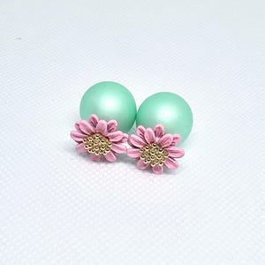 May include: A pair of pink daisy stud earrings with gold-colored centers, next to two mint green spherical beads. The earrings have a delicate, feminine design, perfect for adding a touch of floral charm to any outfit.