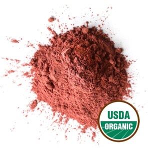 May include: A pile of reddish-brown powder, possibly a cosmetic or food ingredient, is shown against a white background. The powder is coarse and textured. A USDA Organic seal is visible in the lower right corner.