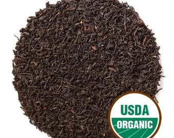 May include: A pile of loose leaf tea, dark brown in color, against a white background. A circular USDA Organic label is visible in the lower right corner. The tea leaves appear to be dried and have a slightly coarse texture.