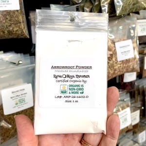 Hand holding a clear plastic zip bag of arrowroot powder. The bag is labeled with the Raven Moon Emporium logo.