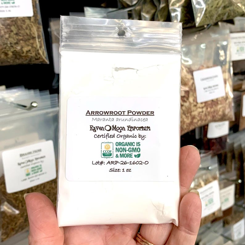 Hand holding a clear plastic zip bag of arrowroot powder. The bag is labeled with the Raven Moon Emporium logo.