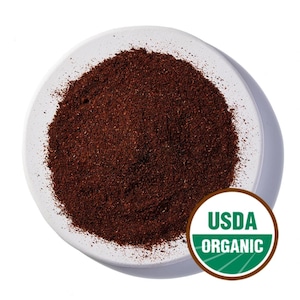 Può includere: Una ciotola di ceramica bianca piena di un mucchio di polvere marrone scuro finemente macinata. La ciotola è posta su uno sfondo bianco. Un sigillo USDA Organic è visibile nell'angolo in basso a destra, a indicare la certificazione biologica del prodotto.