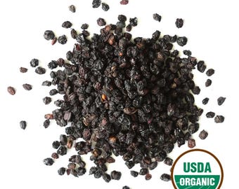 May include: A pile of dried, dark purple elderberries on a white background. The berries are small, shriveled, and have a slightly wrinkled texture. A USDA Organic seal is visible in the lower right corner.