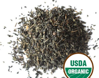 May include: A heap of dark green loose leaf tea against a white backdrop. The tea leaves are scattered. A USDA Organic seal is in the bottom right corner.