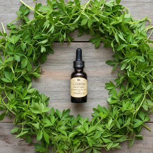 May include: A brown glass bottle of "Oil Cleavers" with a dropper, centered within a wreath of green leaves. The label includes text. The background is a wooden surface.