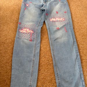 May include: Light blue denim jeans featuring distressed areas repaired with visible pink embroidery stitching and floral patterns. The trousers include a standard waistband with a button closure, belt loops, and a straight-leg cut with hemmed cuffs.