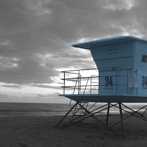May include: A blue lifeguard stand with a black metal frame and a white sign that reads "34 Carlsbad". The stand is on a sandy beach with a gray sky in the background.