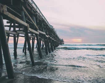 May include: A wooden pier extends out over a blue ocean with white capped waves. The pier is made of dark wood and has many supports. The sky is a light blue with a pink hue.