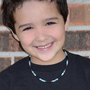 May include: A young boy with short brown hair is smiling and wearing a black t-shirt. He is wearing a beaded necklace with black, gold, and blue beads.