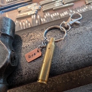 May include: A keyring featuring a brass-coloured metal bullet casing and a small rectangular copper-toned tag stamped with the text Gpa. The keyring includes a silver-toned split ring and a lobster claw clasp, positioned near a metal hammer.
