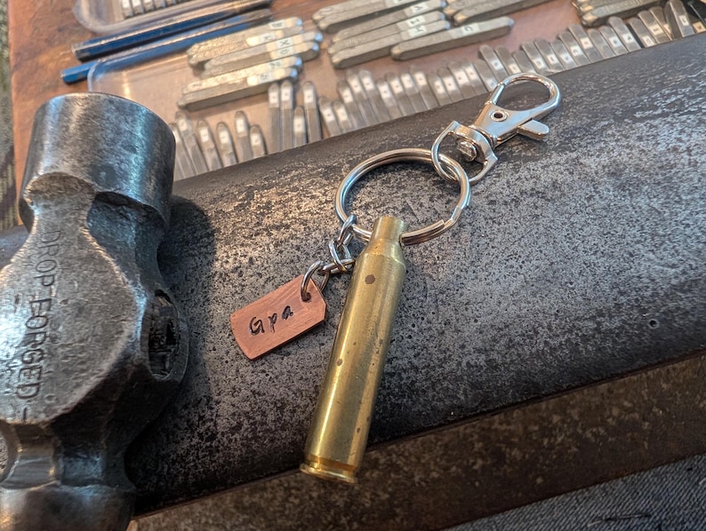 May include: A keyring featuring a brass-coloured metal bullet casing and a small rectangular copper-toned tag stamped with the text Gpa. The keyring includes a silver-toned split ring and a lobster claw clasp, positioned near a metal hammer.