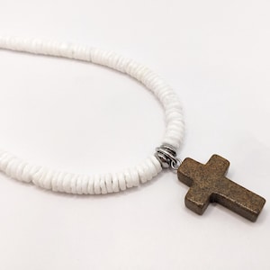 May include: A white beaded necklace with a bronze-colored cross pendant. The cross is a simple, classic design. The necklace is made of small, round white beads.