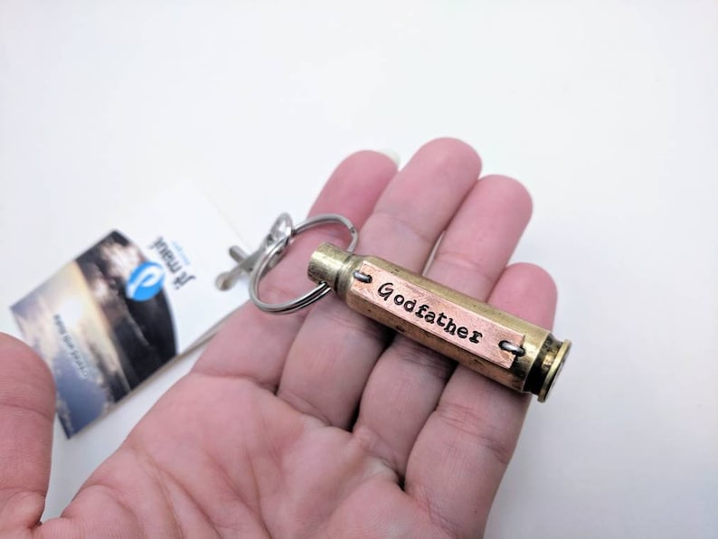 May include: A brass bullet casing keyring featuring a rectangular copper plate attached to the side. The copper plate is stamped with the word Godfather in black lettering. A silver-toned metal split ring is attached to the top of the casing.