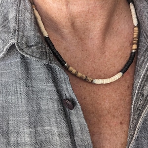 May include: A close-up of a layered beaded necklace with black, white and brown beads. The necklace is worn by a person with a grey shirt.