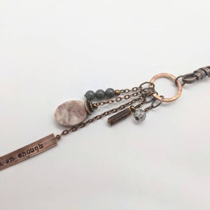 May include: A copper necklace with a charm that says "I am enough" and features a pink and white oval stone, gray beads, and a copper ring.