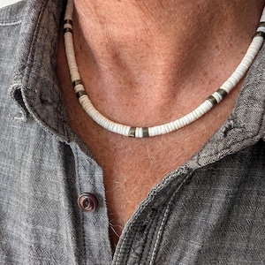 May include: A white beaded necklace with dark metal spacers. The necklace is worn against a grey button-down shirt. The beads are small and round, creating a simple, stylish accessory.