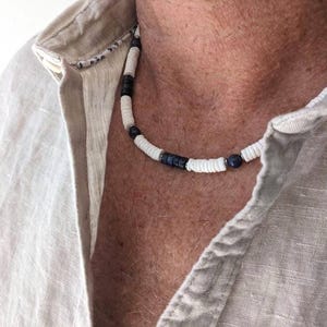 May include: A necklace featuring a repeating pattern of small white disc-shaped beads interspersed with dark blue cylindrical beads and smaller dark blue spherical beads, worn around the neck against a light beige linen-textured collared shirt.