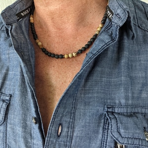 May include: A black and brown beaded necklace with a lava rock and jasper stone design. The necklace is worn by a person wearing a blue denim shirt.