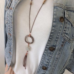 May include: A long necklace featuring a dark metal chain, a textured circular pendant, a pale green rectangular stone, and a brown leather tassel. The necklace is worn over a white ribbed top and a light blue denim jacket with metal buttons.