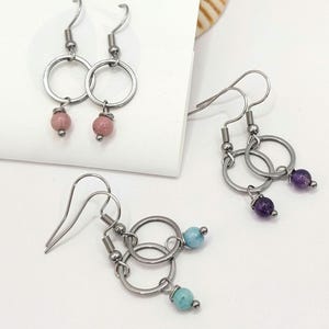 May include: A collection of handmade earrings featuring silver-tone metal hoops and colorful gemstone beads. The earrings include pink, purple, and turquoise beads. The earrings are displayed on a white surface, showcasing their design and craftsmanship.