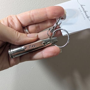 May include: A silver-toned metal bullet casing keyring featuring a small rectangular copper-coloured tag engraved with the name Zeb. The keyring includes a circular split ring and a lobster claw clasp, attached to a white tag reading handmade with aloha.