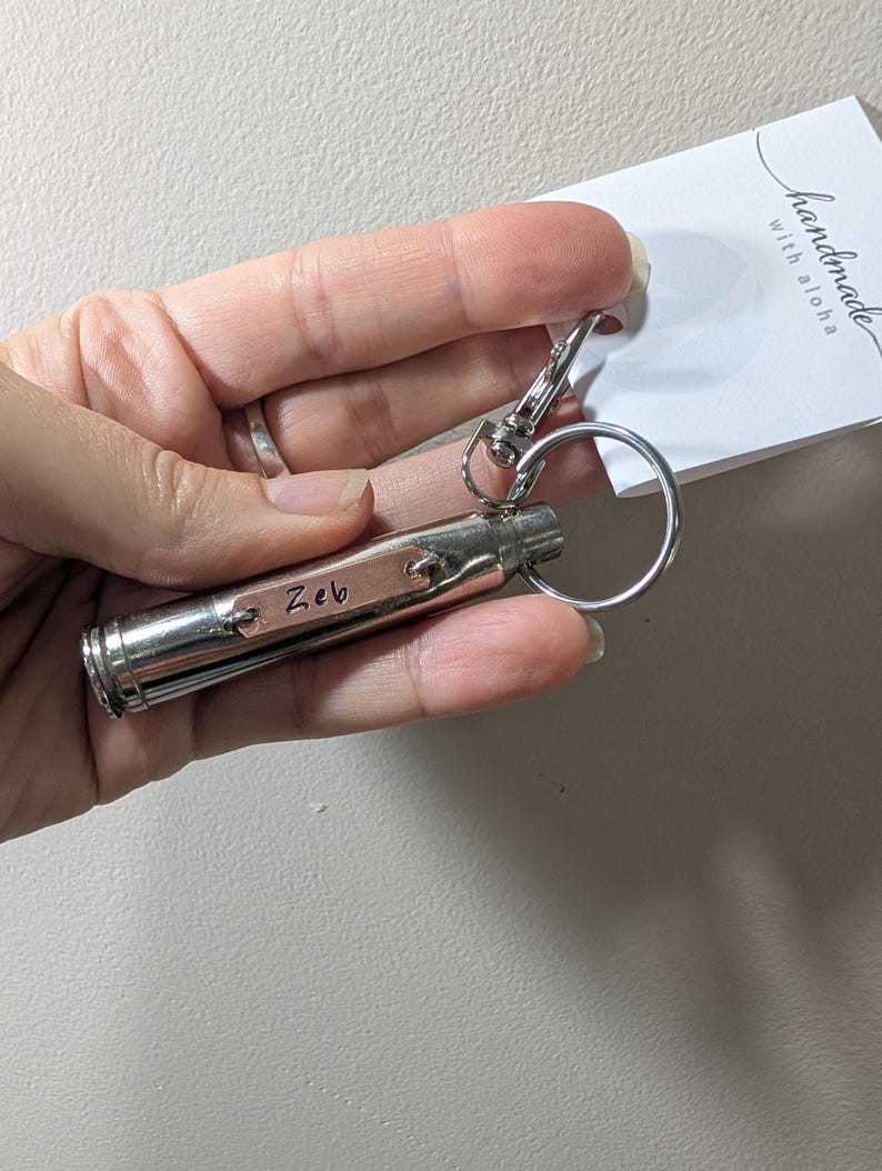May include: A silver-toned metal bullet casing keyring featuring a small rectangular copper-coloured tag engraved with the name Zeb. The keyring includes a circular split ring and a lobster claw clasp, attached to a white tag reading handmade with aloha.