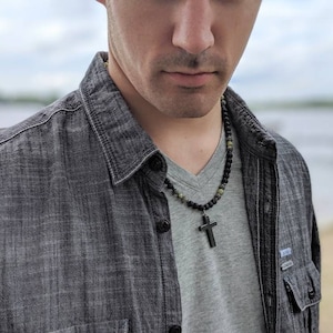 May include: A man wearing a grey shirt and a black and green beaded necklace with a black cross pendant.