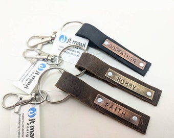 May include: Three leather keychains with metal rings and personalized metal tags. The tags are engraved with the words "GODFATHER", "HOHHY", and "FAITH".