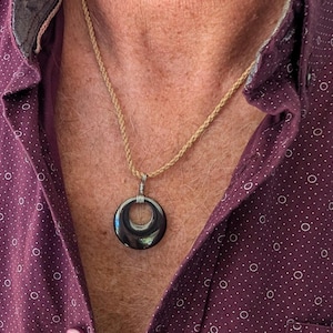May include: A dark, circular pendant necklace with a silver bail hangs on a tan rope chain. The pendant is a dark, glossy color. The necklace is worn over a maroon button-down shirt with a white polka dot pattern.