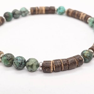 May include: A beaded bracelet featuring alternating dark brown cylindrical beads and round turquoise-colored beads. The bracelet is strung on a silver-colored cord and is displayed against a white background.