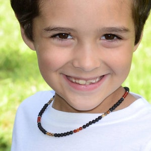 May include: A young boy with short brown hair smiles while wearing a white t-shirt and a black beaded necklace with red and brown accents.