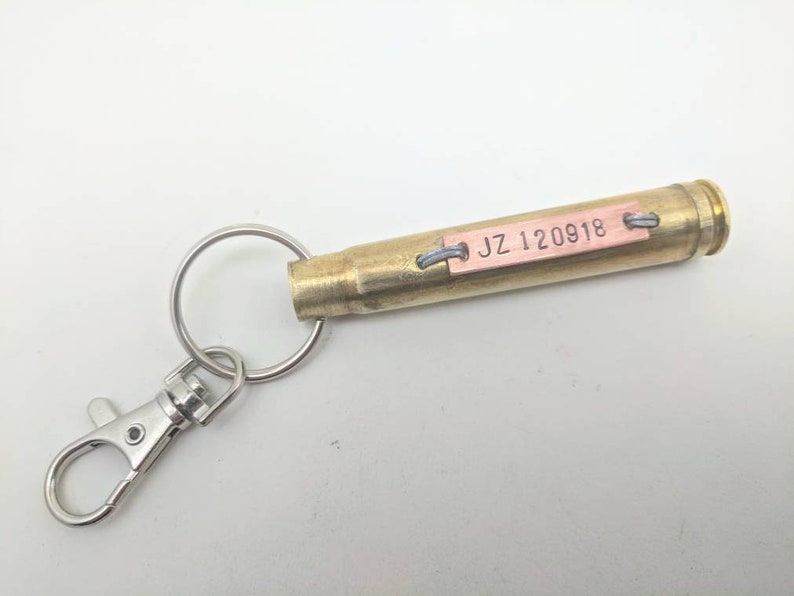 May include: A brass bullet casing keyring featuring a silver-toned metal split ring and lobster clasp. A rectangular copper-coloured metal tag is attached to the side of the casing with wire, stamped with the black text JZ 120918.