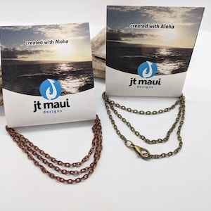 May include: Two chain necklaces, one copper and one bronze, with a lobster clasp closure. Both necklaces are displayed on a wooden surface with a card that reads "created with Aloha" and "jt maui designs".