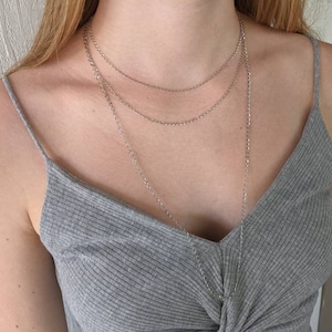 May include: A silver necklace set with three layered chains of varying lengths. The chains are delicate and hang against a gray ribbed tank top. The necklace is a simple, elegant accessory.