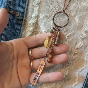 May include: A copper necklace with a charm that says "grace & grit". The charm is attached to a copper circle with a variety of beads and stones, including a large yellow stone.
