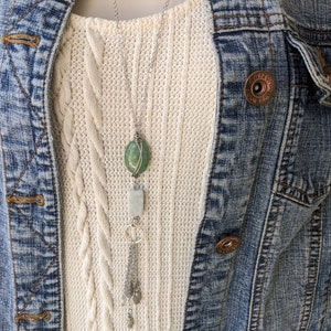May include: A silver chain necklace with a green stone pendant and a silver charm with a tassel. The necklace is layered over a white cable knit sweater.