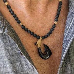 May include: A black hook pendant necklace with a cord, strung with black lava beads and tan-coloured spacers. The necklace is displayed against a grey shirt.
