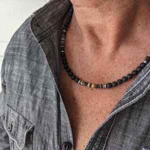 May include: A black lava stone bead necklace with interspersed tan and silver-toned beads. The necklace is worn over a gray denim shirt, creating a casual, earthy aesthetic. The necklace is a stylish accessory.