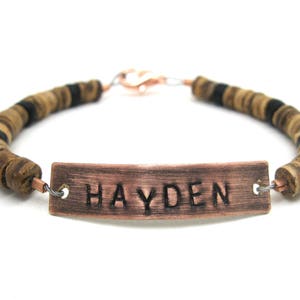 May include: A copper bracelet with a personalized nameplate that reads "HAYDEN". The bracelet is made with brown and black wooden beads.