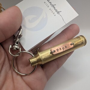 May include: A brass bullet casing keyring featuring a small rectangular copper tag stamped with the word papa. The keyring includes a silver-toned metal split ring and a lobster claw clasp, attached to a white card with the text handmade with aloha.
