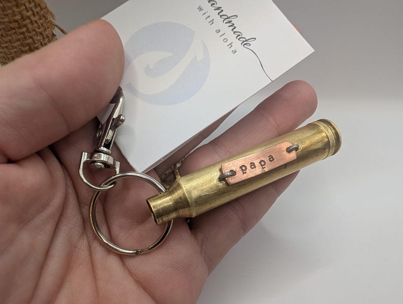 May include: A brass bullet casing keyring featuring a small rectangular copper tag stamped with the word papa. The keyring includes a silver-toned metal split ring and a lobster claw clasp, attached to a white card with the text handmade with aloha.