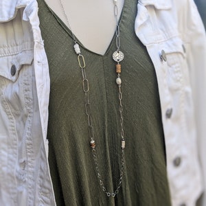 Puede incluir: Un collar largo de cadena plateada con eslabones ovalados y redondos de oro. El collar presenta un colgante de plata con un diseño circular recortado, cuentas de madera y cuentas blancas.