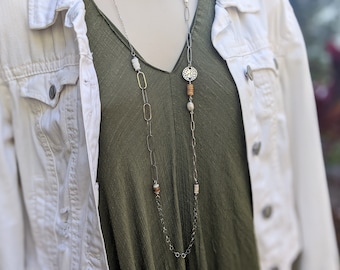May include: A long silver chain necklace with oval and round gold links. The necklace features a silver pendant with a circular cutout design, wooden beads, and white beads.