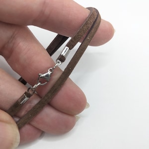 May include: A close-up of a brown suede necklace with a silver-colored clasp. The necklace is held in a hand against a white background. The necklace appears to be a double-strand design.